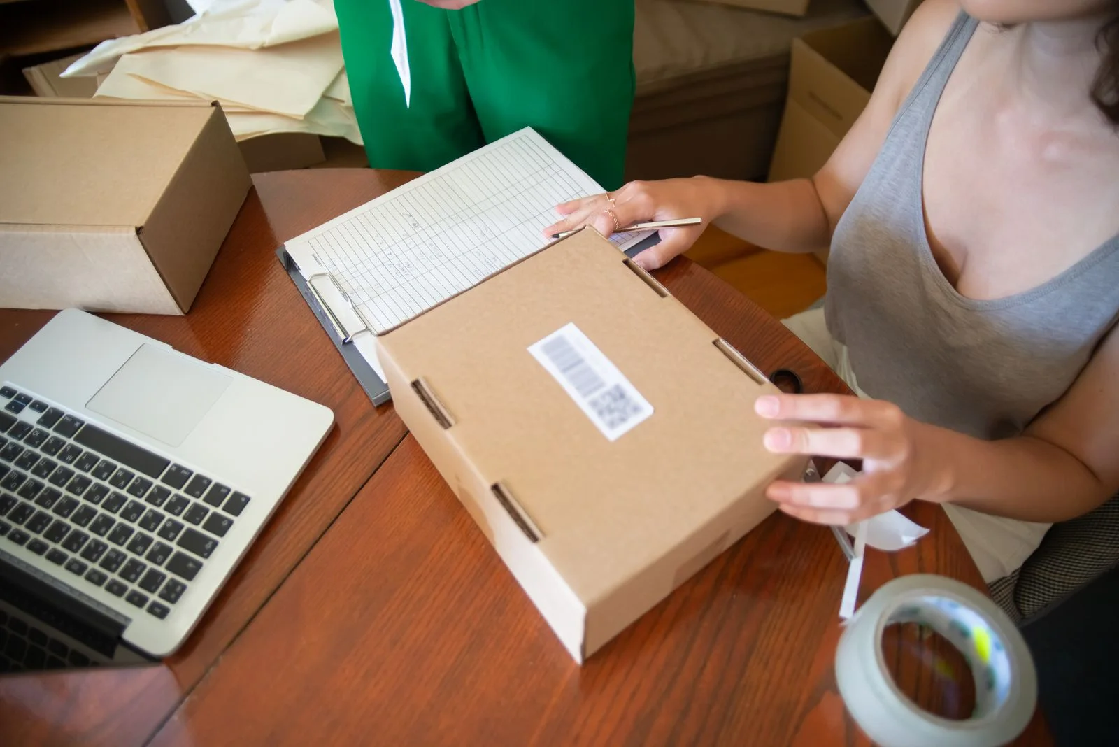 A person prepares a labelled cardboard box on a table beside a laptop and a record sheet.