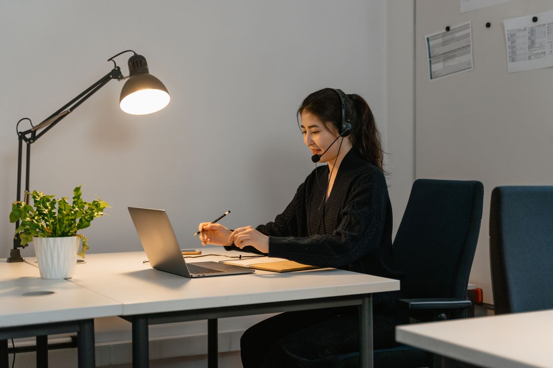 A woman wearing a headset with a microphone works at a laptop, holding a pencil over a notebook.