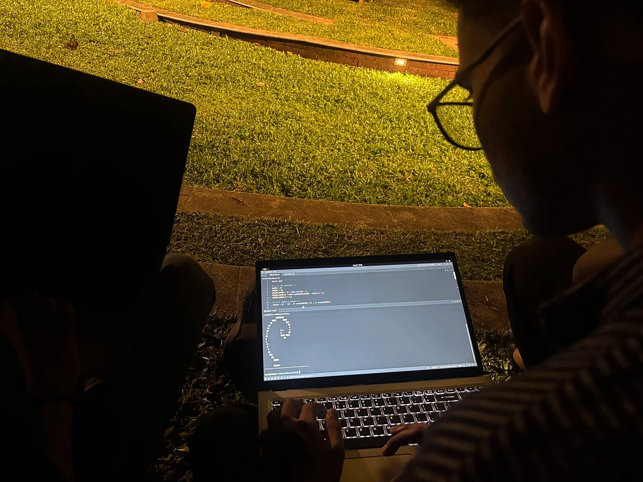 A person wearing glasses working outdoors on a laptop with code on its screen.
