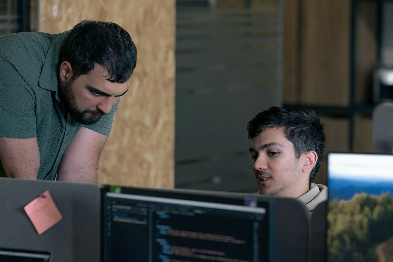 Two people work at several monitors; one is seated and the other leans forward beside them. A screen displaying code is visible in the foreground.